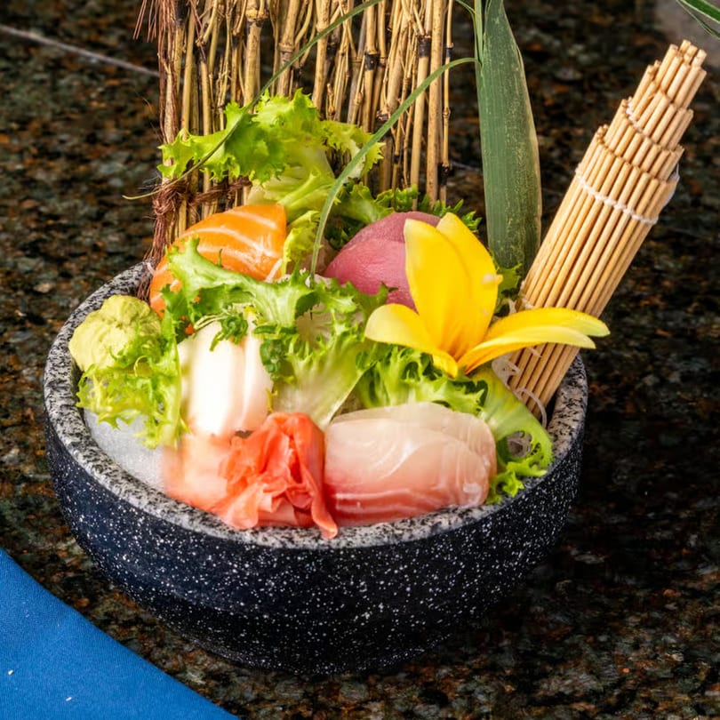 Assorted sashimi with lettuce, wasabi, and ginger in a dark speckled bowl, garnished with a yellow flower.